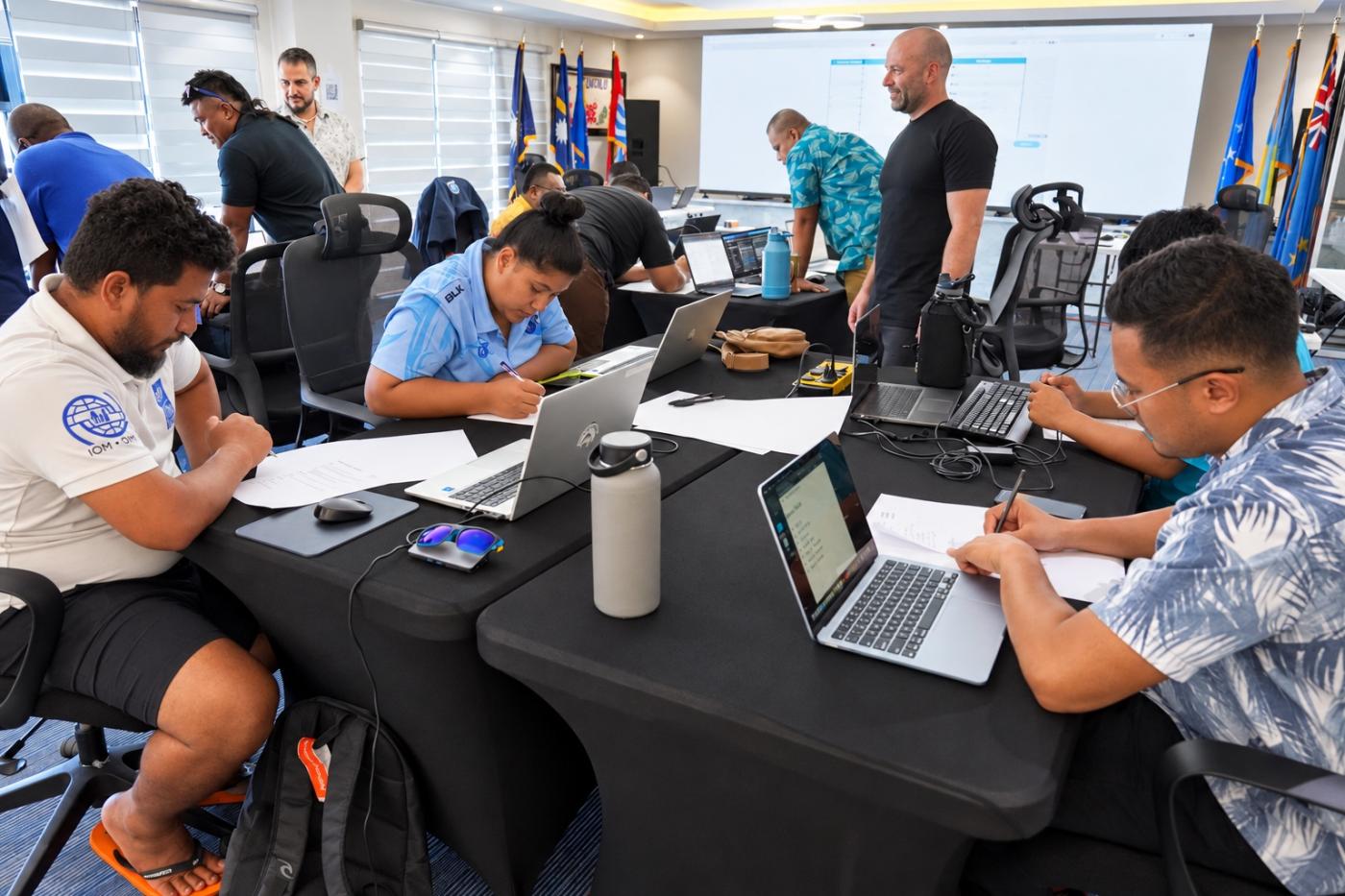 Participants at black-draped tables write notes beside open laptops in a busy training room, with national flags and a large screen behind them.