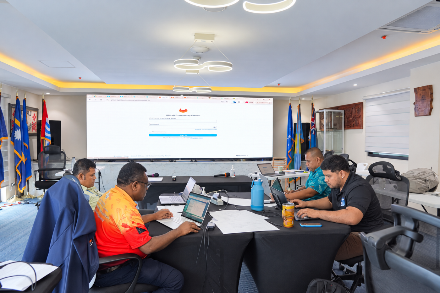 Four participants work on laptops at a black-draped table in a training room, with a GitLab sign-in page on the large screen behind them.