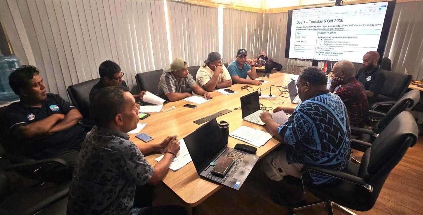 PCCR training in Kiribati: participants around a wooden meeting table with laptops and papers, facing a screen that shows the Day 1 agenda.