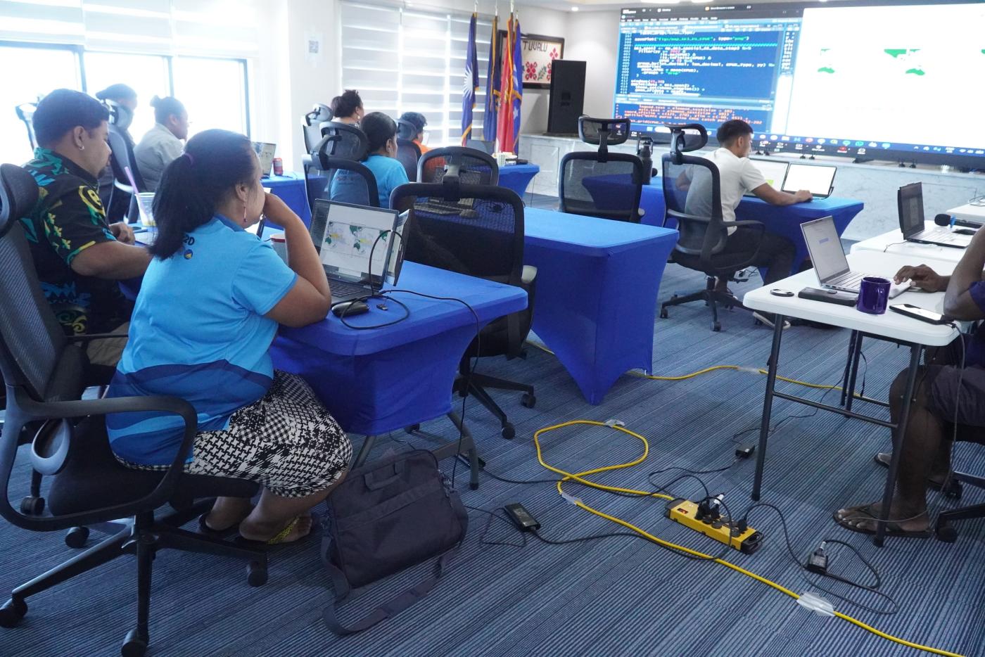 Participants at blue-covered tables with laptops in a training room, watching a large screen that shows programming code and a map.