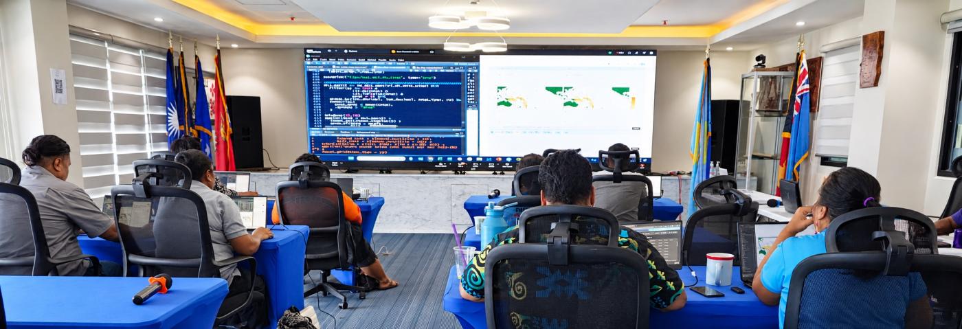Participants seen from behind at blue-covered tables with laptops, facing a wide screen that shows programming code and maps, with national flags on either side.